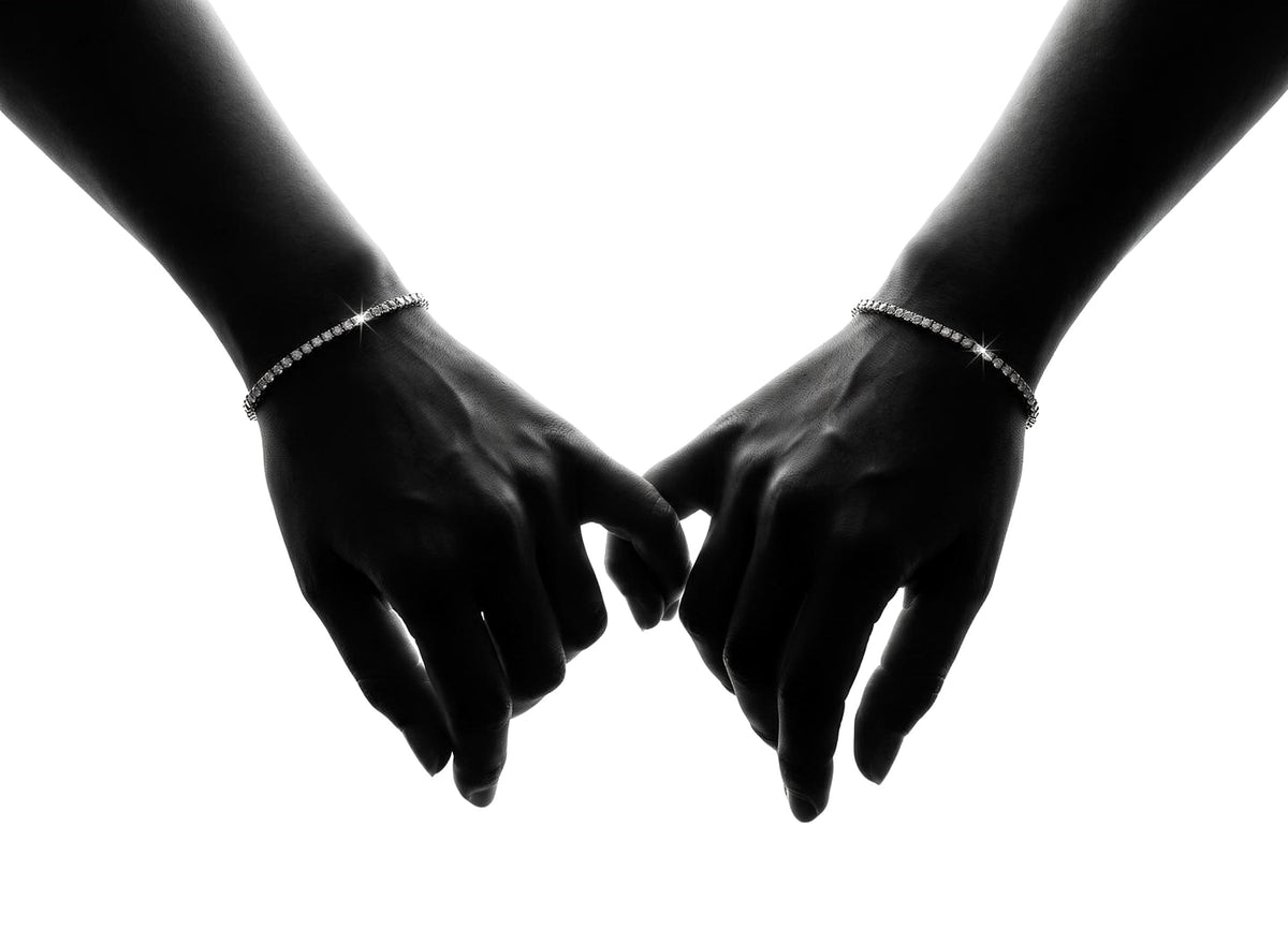 Two hands reaching for each other, each wearing a 2mm lab-grown moissanite tennis bracelet (illustrative render)